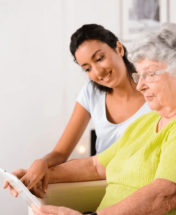A young woman helps an elderly woman use a tablet. Both are seated in a bright, indoor setting.