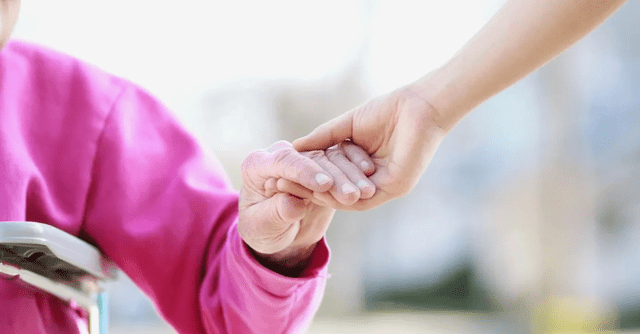 An elderly person in a pink shirt sitting in a wheelchair holds hands with a younger person extending support.