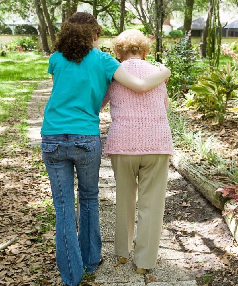 A younger person in a blue shirt supports an older person in pink walking along a park path.