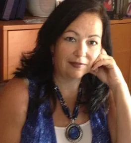 A woman with dark hair, wearing a blue top and necklace, sits with her hand resting on her face in front of a wooden cabinet.