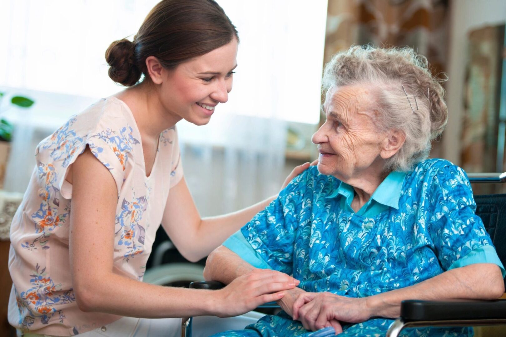 A young woman is smiling and holding hands with an elderly woman in a wheelchair. They are indoors, and the elderly woman is wearing a blue patterned dress.