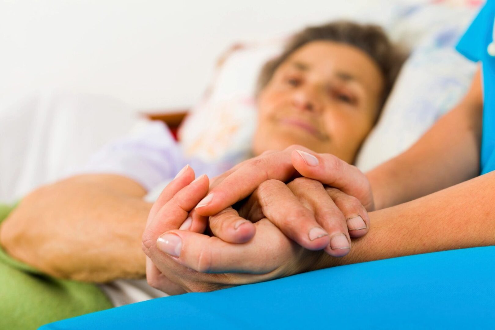 A person in a blue outfit holds the hand of an elderly person lying in bed, offering support and comfort.