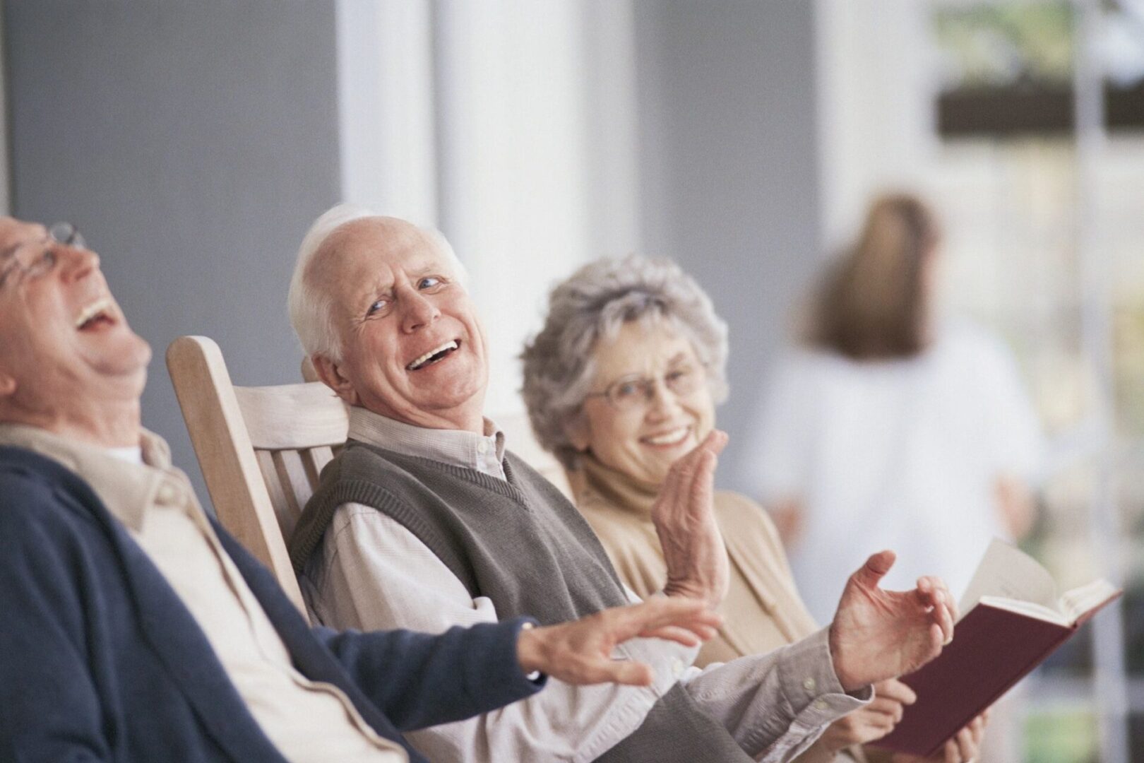 Three older adults are sitting and laughing together. One man in a rocker gestures, while the woman holds a book. A blurred figure stands in the background.