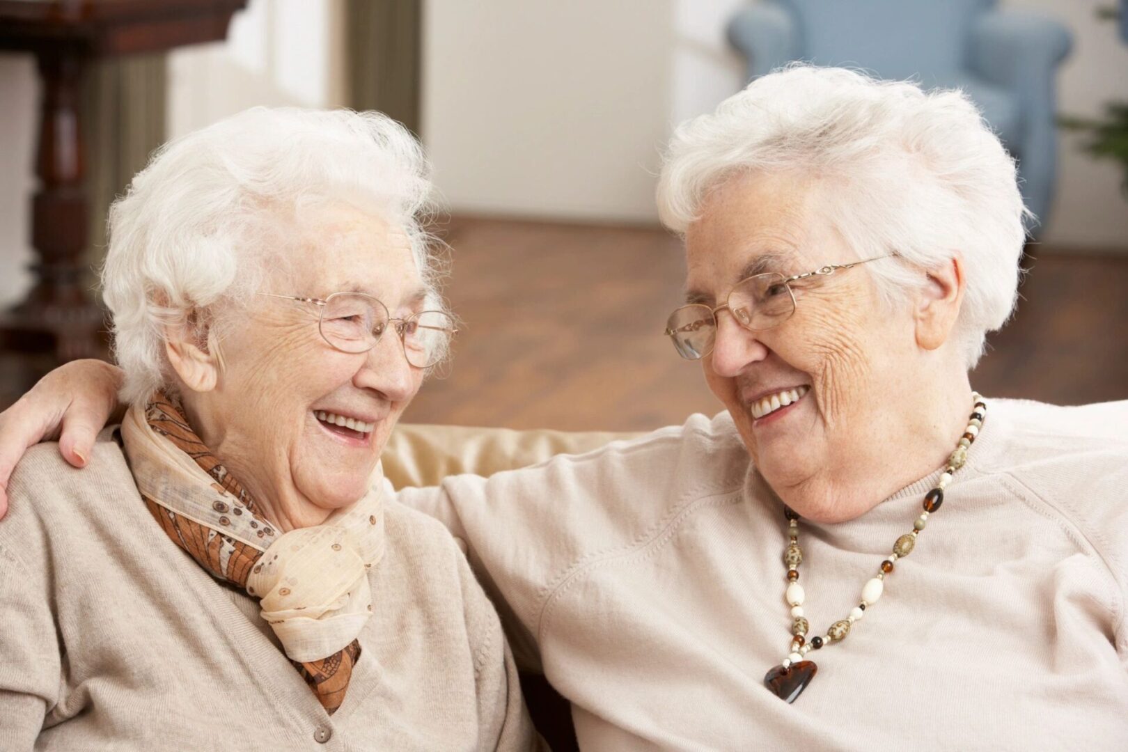 Two elderly women with white hair are sitting closely on a couch, smiling and looking at each other. One has her arm around the other. Both wear glasses and comfortable, light-colored clothing.