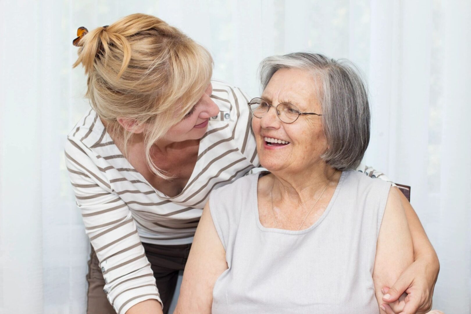 A woman with blonde hair smiles and holds the arm of an elderly woman with gray hair and glasses. Both appear to be enjoying each other's company.