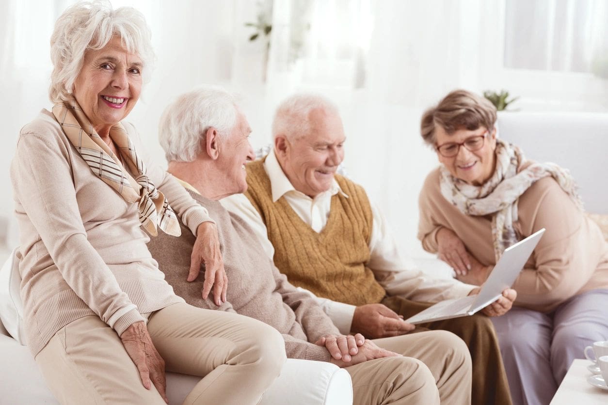 Four elderly individuals are sitting on a couch, three men and one woman, looking at a laptop, with another elderly woman smiling and posing on the armrest.