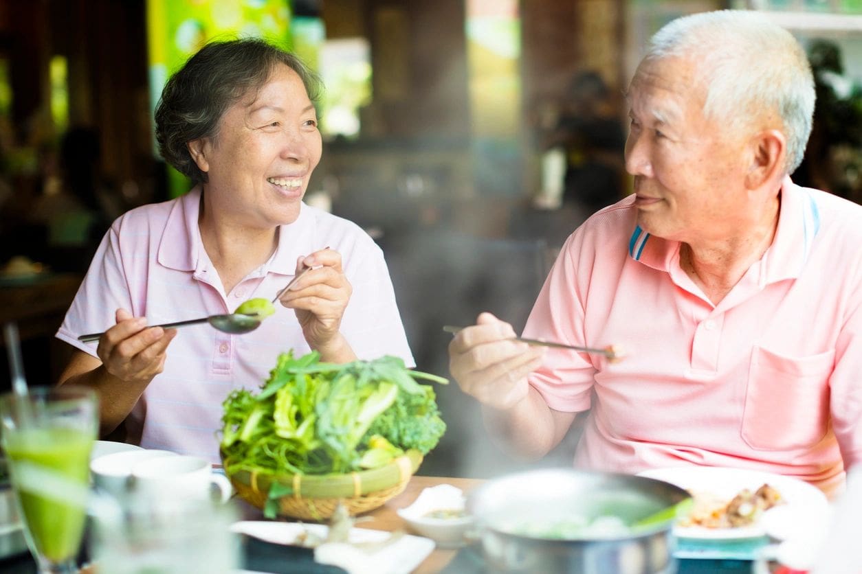 Two elderly individuals sitting at a restaurant table, smiling and eating from a steaming hot pot. A basket of fresh greens and beverages are visible on the table.
